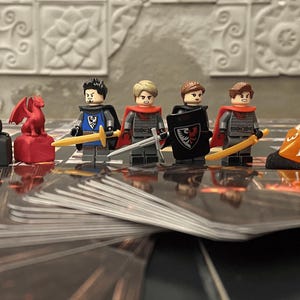 Puede incluir: Una configuraci&oacute;n de juego de mesa con figuras en miniatura y piezas de juego. La escena incluye figuras de estilo Lego de caballeros, un esqueleto y figuras de animales. Tambi&eacute;n se ven dados y cartas de juego, lo que sugiere un tema de fantas&iacute;a o aventura.