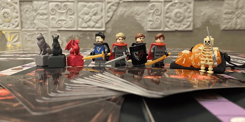 Puede incluir: Una configuraci&oacute;n de juego de mesa con figuras en miniatura y piezas de juego. La escena incluye figuras de estilo Lego de caballeros, un esqueleto y figuras de animales. Tambi&eacute;n se ven dados y cartas de juego, lo que sugiere un tema de fantas&iacute;a o aventura.