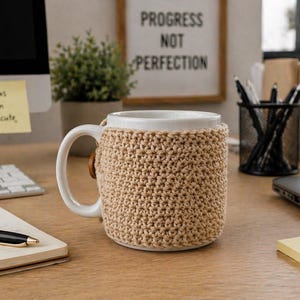 May include: A white ceramic mug with a tan crocheted cozy, sitting on a wooden desk. The mug has a large handle and a wooden button detail on the cozy. A notepad, pen, and sticky notes are also on the desk.