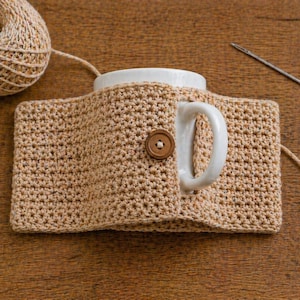 May include: A beige crocheted mug cozy with a wooden button closure, wrapped around a white mug. A ball of beige yarn and a needle are in the background. The cozy is designed to keep beverages warm.