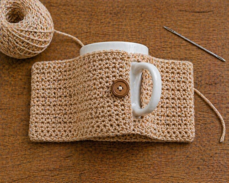 May include: A beige crocheted mug cozy with a wooden button closure, wrapped around a white mug. A ball of beige yarn and a needle are in the background. The cozy is designed to keep beverages warm.