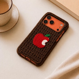 May include: A brown crocheted phone case featuring a red apple with a white bite mark and a green leaf. The case is designed to fit a smartphone, with openings for the camera and buttons. The case is on a white surface.