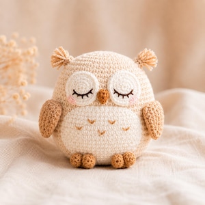 Puede incluir: Una figura de búho de ganchillo en beige y crema. El búho tiene los ojos cerrados con largas pestañas, mejillas rosadas y pequeñas alas texturizadas. Los pies y los mechones de las orejas del búho son de un beige más oscuro. El búho está sobre una tela color crema.