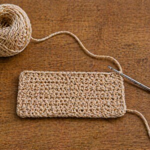 May include: A ball of beige yarn with a crocheted rectangle and a crochet hook on a wooden surface. The crochet project is in progress, with the hook inserted into the stitches. The yarn is a neutral color, suitable for various craft projects.