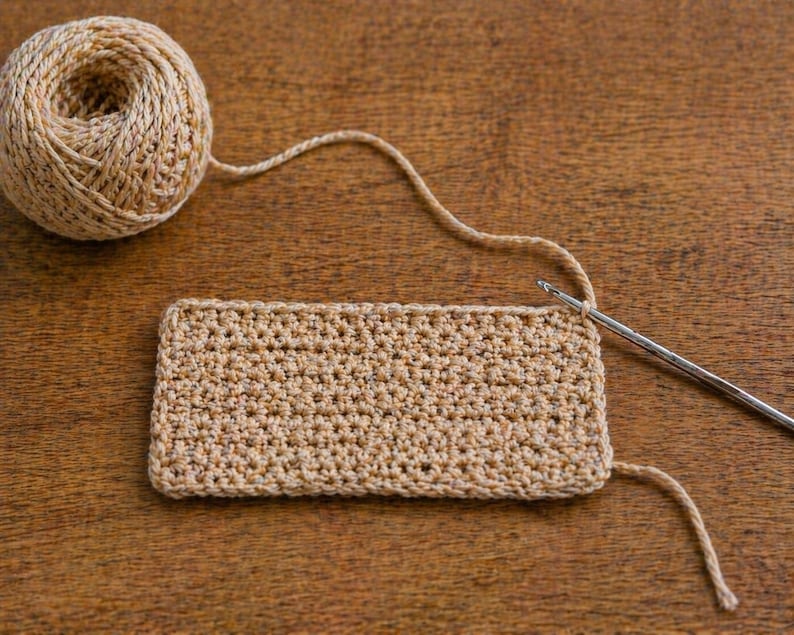 May include: A ball of beige yarn with a crocheted rectangle and a crochet hook on a wooden surface. The crochet project is in progress, with the hook inserted into the stitches. The yarn is a neutral color, suitable for various craft projects.