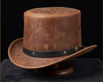 May include: A brown leather top hat with a black leather band and buckle. The hat has a wide brim and is displayed on a wooden stand. The hat has four small holes on the side.