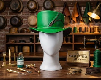 May include: A bright green leather top hat with a dark brown zipper detail along the side, displayed on a white mannequin head. The hat features a structured crown and a curved brim, showcasing a smooth finish and distinct zipper embellishment.