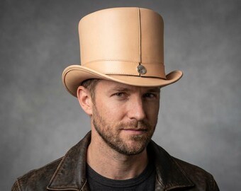 May include: A light beige leather top hat featuring a structured crown, a curved brim, and a matching leather band around the base. A small, circular metallic emblem is centered on the front of the band, securing the leather strap in place.