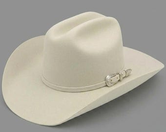 May include: A cream-colored felt cowboy hat with a cattleman crease crown and a wide, curved brim. The hat features a matching cream-colored hat band adorned with a decorative, engraved silver-tone metal buckle set on the side of the crown.