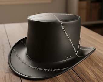 May include: A black leather top hat with a wide brim. The hat features white stitching details along the seams and brim. The hat is sitting on a wooden surface.