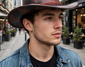 May include: A wide-brimmed hat featuring a dark brown and black mottled pattern with a smooth finish. The hat is accented by a thin, braided reddish-brown cord wrapped around the base of the crown, providing a distinct contrast to the dark tones.