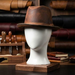 Könnte beinhalten: Ein brauner Leder-Fedora-Hut mit strukturierter Krone und breiter Krempe, präsentiert auf einem weißen Schaufensterpuppenkopf. Der Hut hat eine glatte, matte Oberfläche mit subtilen Farbvariationen und ruht auf einem quadratischen Holzsockel.
