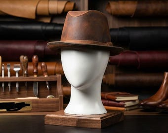 May include: A brown leather fedora hat with a structured crown and a wide brim, displayed on a white mannequin head. The hat features a smooth, matte finish with subtle variations in color, resting on a square wooden base in a workshop setting.