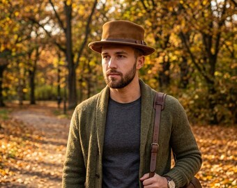 May include: A brown felt fedora hat with a leather band, a charcoal gray t-shirt, and an olive green knit shawl-collar cardigan. The outfit includes a brown leather bag strap worn over the shoulder and a wristwatch with a brown leather band.