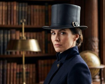 May include: A black top hat featuring a structured crown, a flat brim, and a metallic buckle detail on the side band. The hat is constructed with visible vertical seams and a smooth, matte finish, providing a classic silhouette for formal headwear.