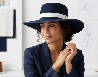 May include: A navy blue wide-brimmed felt hat featuring a structured crown and a contrasting white ribbon band encircling the base. The hat has a smooth, matte finish and a wide, slightly downturned brim that provides a clean, uniform appearance.