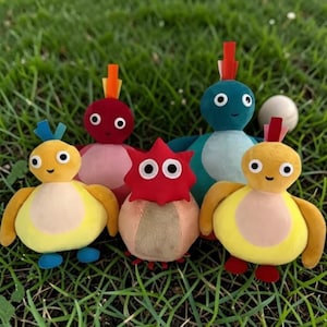 May include: A group of soft toys in various colours, including yellow, red, blue, and pink. Each toy has a distinct design, with large, round eyes. They are arranged on a bed of green grass, creating a playful scene.