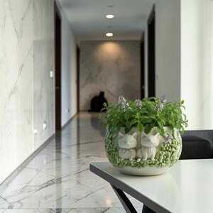 Puede incluir: Una maceta de cerámica blanca con un diseño en relieve verde de dos gatos y follaje, llena de plantas verdes y flores moradas. La maceta está sobre una mesa blanca en un pasillo moderno con paredes de mármol.
