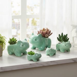 Peut inclure: Collection de cinq jardinières en céramique vert clair en forme de personnage de dessin animé. Chaque jardinière contient une plante succulente. Les jardinières sont de tailles variées et sont disposées sur une surface blanche près d'une fenêtre.