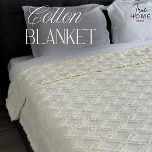 Może przedstawiać: Kremowy koc bawełniany z wypukłym wzorem w romby i postrzępionymi krawędziami. W górnej części widoczny jest napis Cotton BLANKET oraz True HOME STORE. Koc jest rozłożony na łóżku z białymi poduszkami.