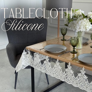 May include: A clear silicone tablecloth featuring an intricate white lace border pattern draped over a wooden dining table. The table is set with grey plates, green textured glasses, and a vase of white flowers, with the text TABLECLOTH Silicone.
