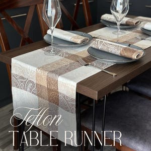 Op de afbeelding: Een bruine houten eettafel met een Teflon tafelloper. De tafelloper heeft een paisley-ontwerp in bruin- en crèmetinten. De tafel is gedekt met borden, servetten en wijnglazen. De tekst "Teflon TABLE RUNNER" is zichtbaar.