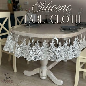 Könnte beinhalten: Eine transparente Silikon-Tischdecke mit weißer Spitzenborte, die einen runden Holztisch bedeckt. Auf der Tischdecke steht in weißer Schrift "Silicone TABLECLOTH". Der Tisch ist mit grauen Tellern und Gläsern gedeckt.