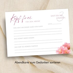 May include: A white card with the words "Kopf frei" in a script font, with prompts for reflection. The card includes the text "FÜR DEN ABEND" and "SANFT DEN TAG LOSLASSEN". Pink flowers are in the lower right corner. The card is on a beige surface.