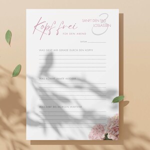 May include: A white journal page with German text and lines for writing. The top of the page has the words "Kopf frei" in a cursive font. The page is decorated with pink flowers and green leaves.