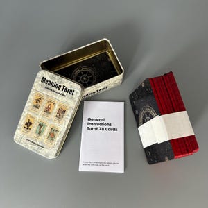 Könnte beinhalten: Ein Tarotkartenset mit einer Metallbox mit der Aufschrift "Meaning Tarot". Das Set enthält 78 Karten, ein Anleitungsheft und eine Metallbox mit goldfarbenem Innenraum. Die Karten sind mit einem weißen Papierband gebunden.