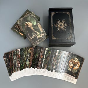 May include: A deck of "Celtic Magic Oracle Cards" with a black box featuring a gold geometric design. The card deck has a woman with long hair on the front. The cards are spread out, showing detailed illustrations and text about lunar cycles.