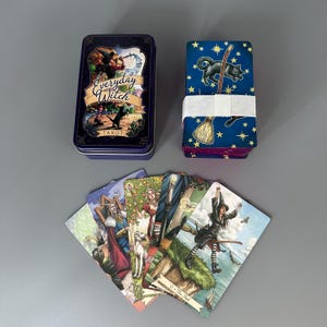 May include: An Everyday Witch Tarot card set. The set includes a blue metal box with the words "Everyday Witch Tarot" and a stack of cards wrapped in white paper. Several tarot cards are fanned out, displaying colourful illustrations.