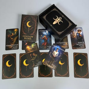 May include: A black tarot card deck and box. The cards display illustrations and text, such as "Shadow Work," "Never Enough," and "Fear of Intimacy." The box has a gold design on the lid.
