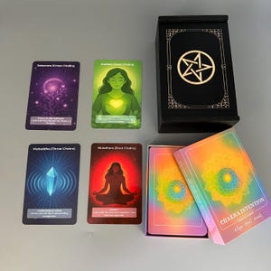 May include: A Chakra Intention Oracle Cards set with a black wooden box featuring a pentagram design. The set includes cards with illustrations and text, such as "Sahasrara (Crown Chakra)" and "Anahata (Heart Chakra)." The box and card packaging have a colorful, rainbow design.