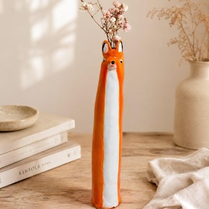 May include: A tall, slender ceramic vase shaped like a fox, featuring a bright orange exterior with a white vertical stripe down the front. The top of the vase depicts a fox head with pointed ears, black eyes, and a small nose, holding dried flowers.