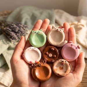 Może przedstawiać: Kolekcja siedmiu małych, okrągłych ceramicznych podstawek w kształcie różnych zwierząt, w tym zielonego kota, różowej świnki, brązowego niedźwiedzia i beżowego psa, z których każda ma gładki, wklęsły środek do przechowywania drobiazgów.