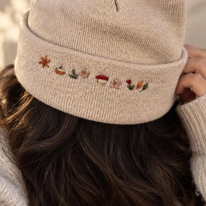 Könnte beinhalten: Eine beige Strickmütze mit umgeschlagenem Saum, der mit kleinen, feinen Stickereien verziert ist, darunter Herbstblätter, ein Kürbis, Blumen und ein Pilz in verschiedenen Farben wie Rot, Orange, Grün und Rosa, mittig auf der Vorderseite.
