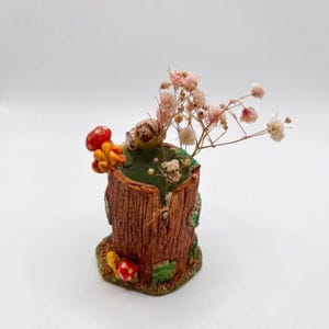 May include: A brown cylindrical vase textured to resemble a tree stump, featuring small sculpted red and yellow mushrooms and green moss accents. The vase contains a cluster of delicate, light pink dried baby's breath flowers.