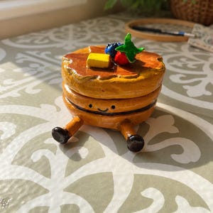 Puede incluir: Un recipiente redondo con forma de tortita dorada y tapa. La parte superior presenta un cuadrado de mantequilla amarillo, un arándano azul y una fresa roja con hojas verdes. La base tiene una pequeña cara sonriente pintada a mano y dos piernas.