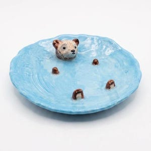 May include: A round, light blue ceramic dish with a textured, wavy surface. A small, sculpted brown bear head and four brown bear paws emerge from the centre, creating the appearance of a bear swimming in water with a glossy, reflective finish.