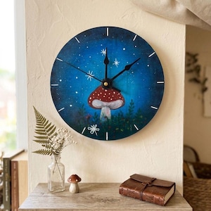 May include: Round wall clock with a hand-painted design featuring a red and white mushroom against a blue starry sky. The clock has black hands and white hour markers. A whimsical piece of home decor.