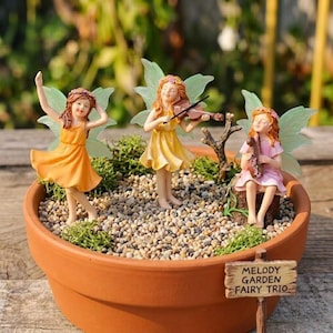 May include: A terracotta plant pot containing a miniature garden scene with three fairy figurines. One fairy dances, another plays a violin, and the third plays a small instrument. The fairies wear colourful dresses and have translucent wings. A sign reads "Melody Garden Fairy Trio."