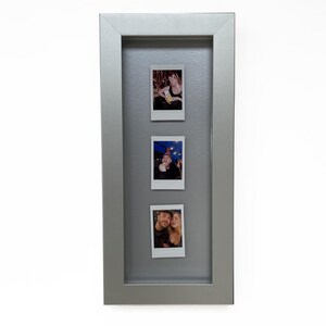 Peut inclure: Un cadre photo rectangulaire argenté au design en boîte profonde contenant trois photographies instantanées verticales disposées en colonne. Le cadre présente une finition métallique lisse et un passe-partout gris entourant les photos.