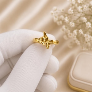 May include: A gold-colored ring with an abstract design. The ring features a textured, organic shape in the center, with a smooth band. The ring is held by a gloved hand against a soft, neutral background.