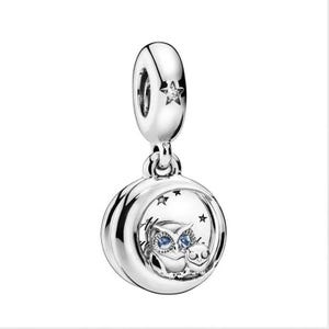 May include: A silver charm featuring a circular frame with a moon and star motif. Inside the circle, two owls are depicted with blue stone eyes. The top bail is adorned with a small star containing a clear stone. The metal has a polished finish.