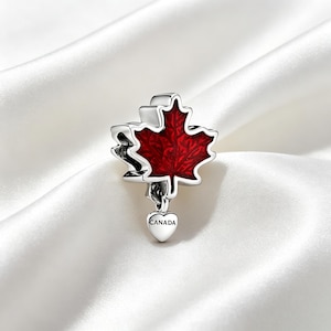 Peut inclure: Un charm en argent en forme de feuille d'érable avec une finition en émail rouge profond. Un petit pendentif en argent en forme de cœur est suspendu au bas de la feuille, avec le mot CANADA gravé en lettres noires sur sa face avant.