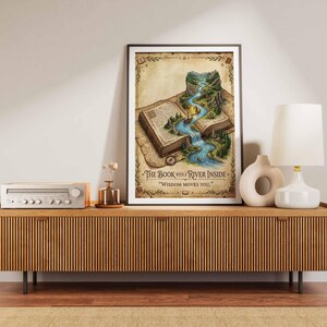 Peut inclure: Une affiche verticale encadrée illustrant un livre ouvert d'où s'écoule une rivière. Le texte indique THE BOOK WITH A RIVER INSIDE et WISDOM MOVES YOU. L'illustration représente un paysage avec des montagnes, des arbres et un petit bateau.