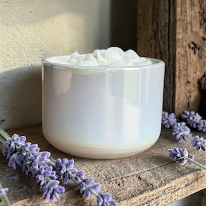 May include: A white, pearlescent candle in a round ceramic pot. The candle has a floral design on top. It sits on a wooden shelf with sprigs of lavender, creating a calming aesthetic.