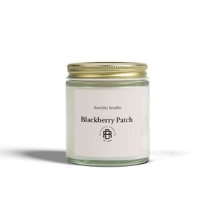 May include: A glass jar candle with a gold lid. The label reads "Ranchito Bendito Blackberry Patch" with a logo. The candle is white and appears to be scented. The jar is clear, allowing the candle's color to be visible.