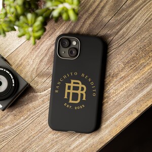 Puede incluir: Funda de teléfono negra con un logotipo dorado que dice "RANCHITO BENDITO EST. 2025". La funda está sobre una superficie de madera, con una cámara y vegetación en el fondo.
